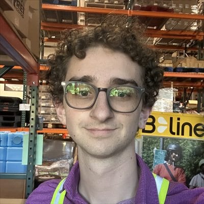 Ari Grayzel, founder of PDX Quake Prep, in a safety vest at the fulfillment warehouse, with kit water containers and pallet racking behind him